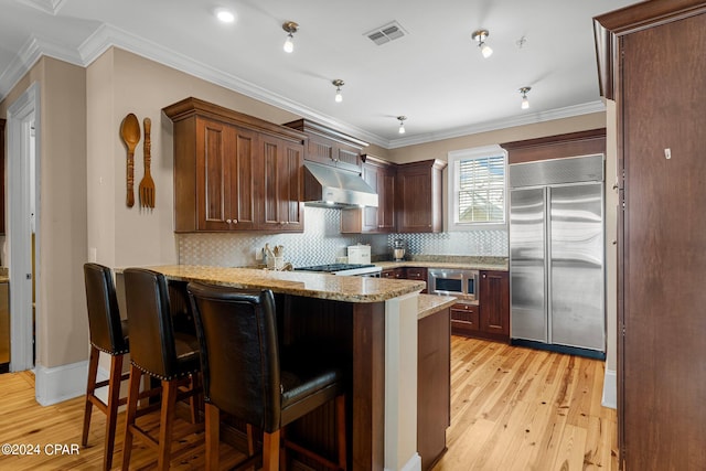 kitchen with kitchen peninsula, built in appliances, light wood-type flooring, crown molding, and a kitchen bar