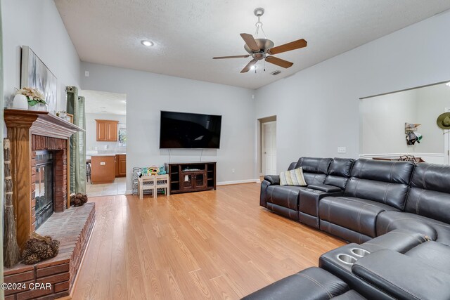 living room with a brick fireplace, ceiling fan, light wood-type flooring, and a textured ceiling