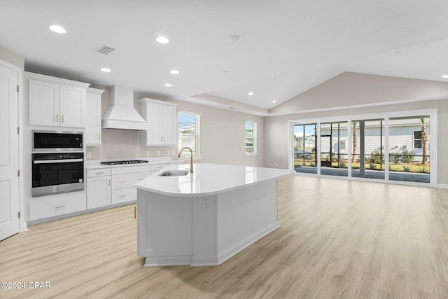 kitchen featuring a kitchen island with sink, appliances with stainless steel finishes, a sink, premium range hood, and light wood-style flooring