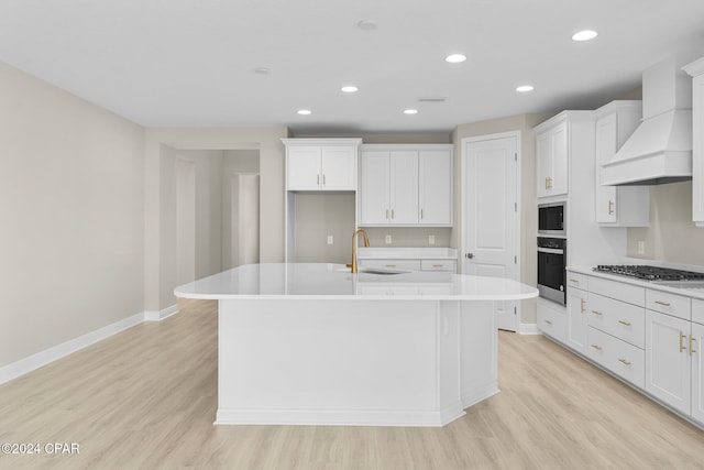 kitchen with stainless steel appliances, an island with sink, custom exhaust hood, and light wood finished floors
