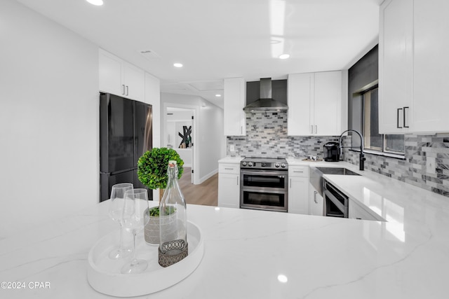 kitchen featuring black refrigerator, white cabinets, range with two ovens, light stone counters, and wall chimney range hood