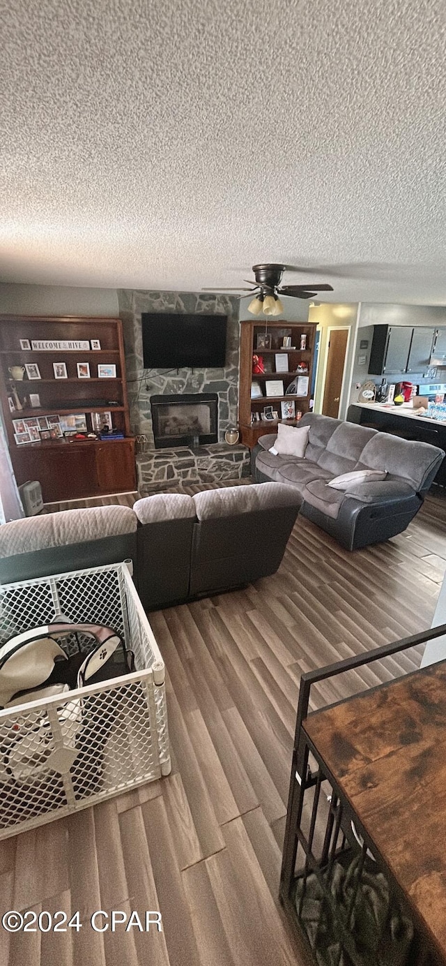 living room featuring wood-type flooring, a textured ceiling, a stone fireplace, and ceiling fan