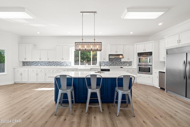 kitchen with a kitchen island with sink, light wood-type flooring, stainless steel appliances, hanging light fixtures, and white cabinetry