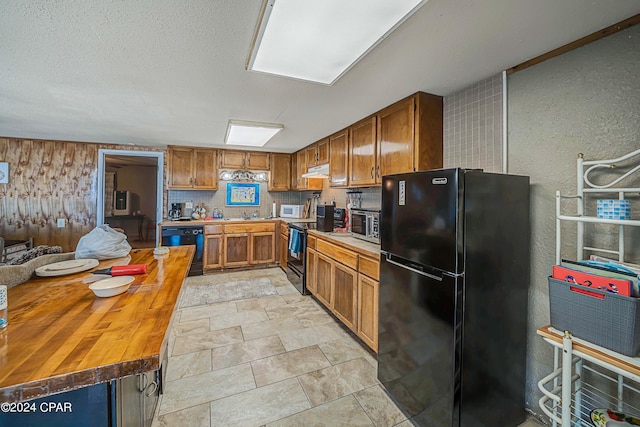 kitchen featuring a textured wall, butcher block counters, brown cabinets, under cabinet range hood, and black appliances