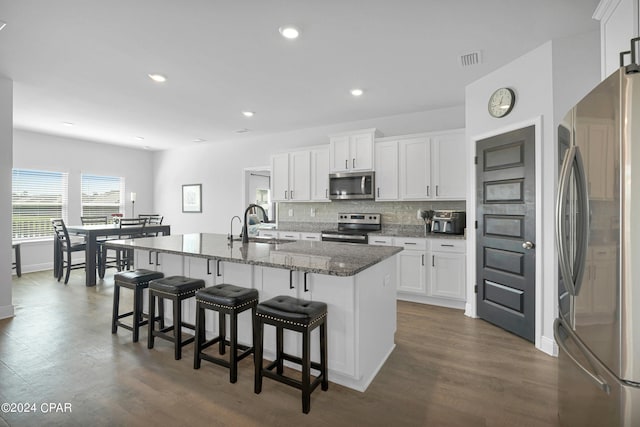 kitchen with stainless steel appliances, dark hardwood / wood-style flooring, white cabinets, dark stone countertops, and a kitchen island with sink