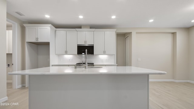 kitchen with light wood-type flooring, backsplash, sink, white cabinets, and an island with sink