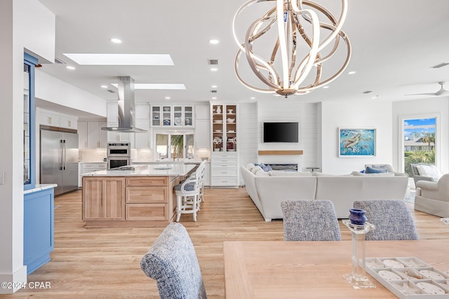 kitchen with a large island with sink, wall chimney range hood, light wood-type flooring, white cabinetry, and stainless steel appliances