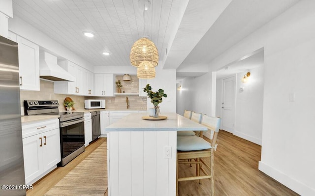 kitchen with appliances with stainless steel finishes, light wood-type flooring, white cabinetry, pendant lighting, and custom exhaust hood