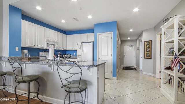 kitchen featuring a peninsula, white appliances, white cabinetry, a kitchen breakfast bar, and dark stone countertops