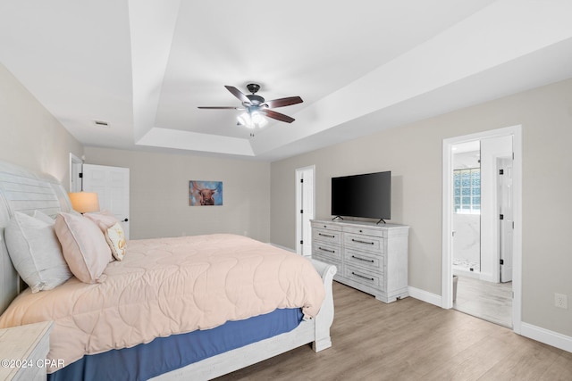 bedroom with ensuite bathroom, a tray ceiling, light wood-type flooring, and ceiling fan