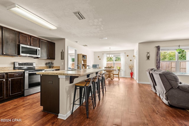 kitchen with a kitchen breakfast bar, dark hardwood / wood-style flooring, stainless steel appliances, a center island with sink, and hanging light fixtures