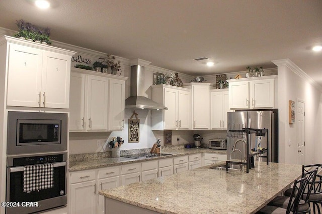 kitchen with white cabinetry, stainless steel appliances, wall chimney range hood, an island with sink, and ornamental molding