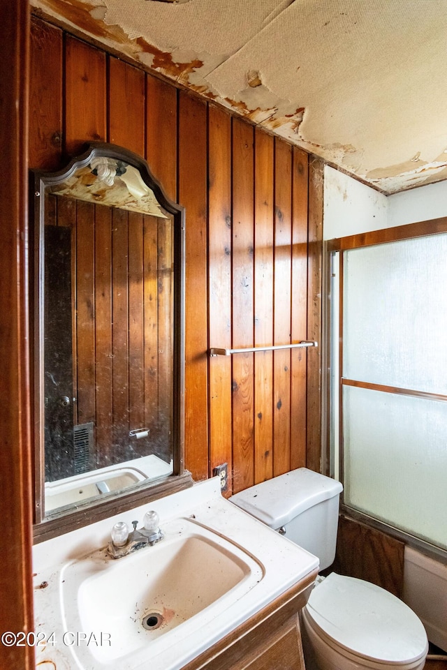 full bathroom with bath / shower combo with glass door, vanity, wooden walls, and toilet