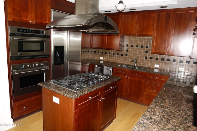 kitchen featuring sink, dark stone countertops, island exhaust hood, stainless steel appliances, and light hardwood / wood-style flooring