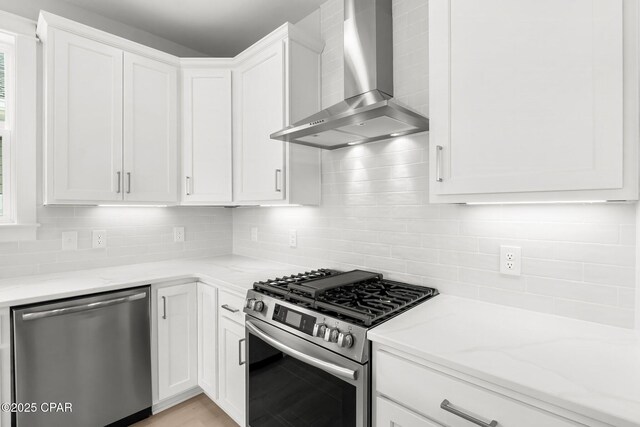 kitchen featuring light stone counters, wall chimney range hood, stainless steel appliances, decorative backsplash, and white cabinets