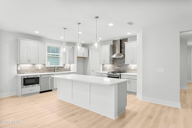 kitchen with appliances with stainless steel finishes, white cabinetry, hanging light fixtures, a center island, and wall chimney range hood