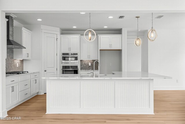 kitchen with hanging light fixtures, stainless steel appliances, a center island with sink, and wall chimney exhaust hood