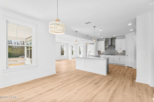 kitchen with decorative light fixtures, tasteful backsplash, white cabinets, wall chimney range hood, and a center island with sink