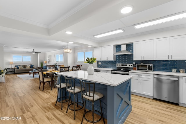 kitchen featuring white cabinetry, a center island, stainless steel appliances, wall chimney range hood, and ceiling fan with notable chandelier