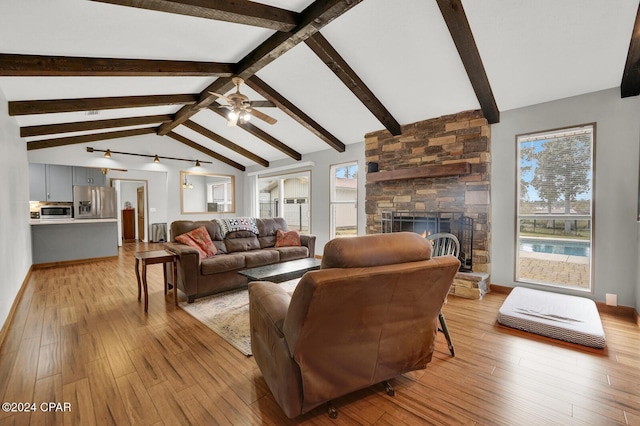 living room with ceiling fan, a stone fireplace, light hardwood / wood-style floors, and lofted ceiling with beams