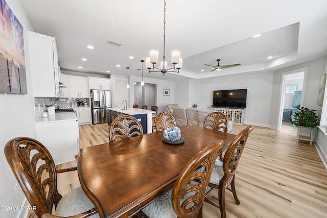 dining room featuring light wood-type flooring, ceiling fan with notable chandelier, sink, and a raised ceiling