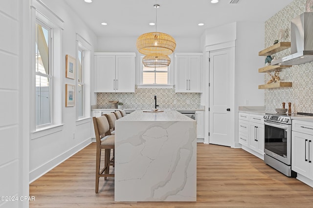 kitchen with pendant lighting, white cabinets, stainless steel range with electric cooktop, wall chimney exhaust hood, and light hardwood / wood-style flooring