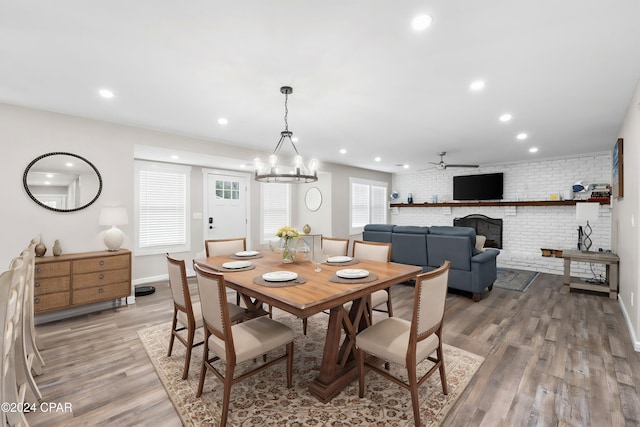 dining area with brick wall, a brick fireplace, ceiling fan with notable chandelier, and light hardwood / wood-style flooring