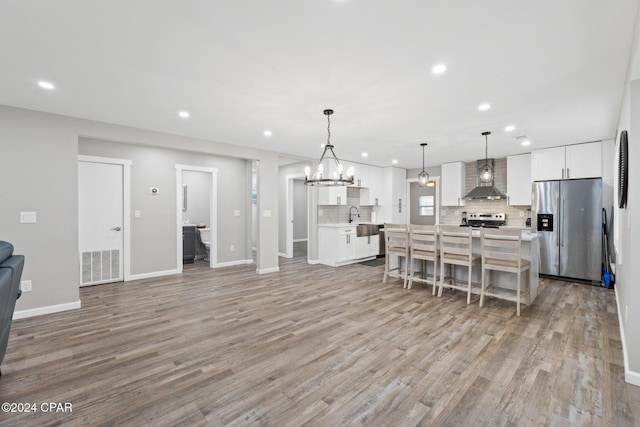 kitchen with pendant lighting, wall chimney range hood, appliances with stainless steel finishes, an island with sink, and white cabinets