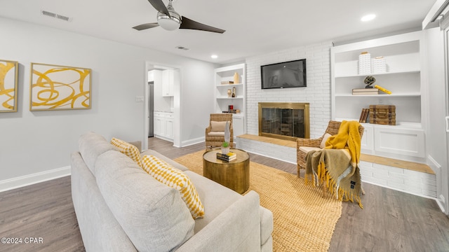 living room with ceiling fan, built in features, dark wood-type flooring, and a brick fireplace