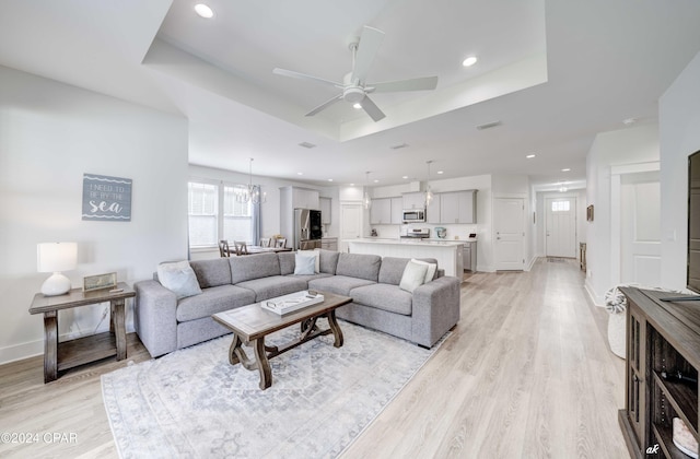 living room featuring ceiling fan with notable chandelier, light wood-type flooring, and a tray ceiling