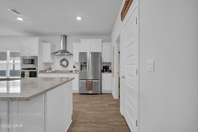 kitchen featuring white cabinetry, stainless steel appliances, light stone countertops, wall chimney range hood, and light wood-type flooring