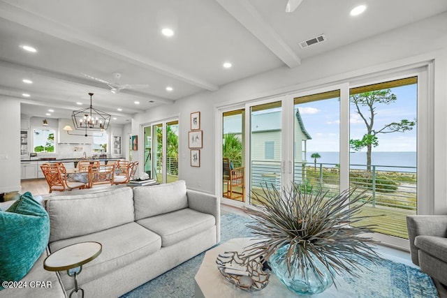 living room featuring beam ceiling, ceiling fan with notable chandelier, a water view, and a wealth of natural light