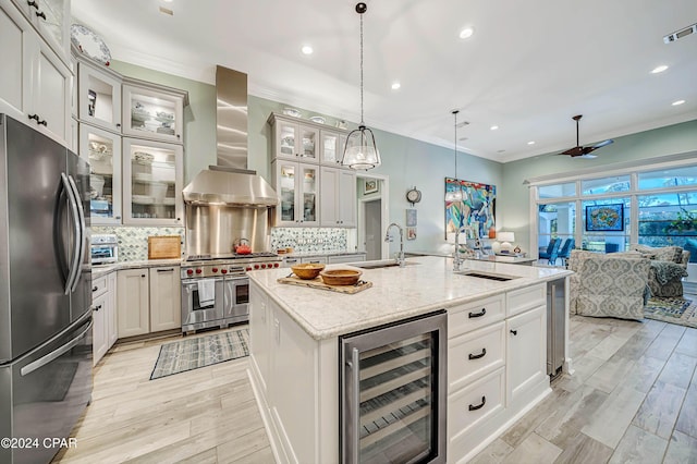 kitchen with sink, appliances with stainless steel finishes, wine cooler, an island with sink, and wall chimney exhaust hood