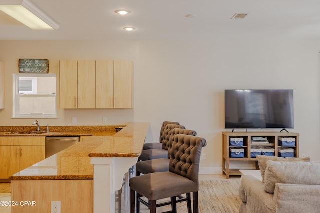 kitchen featuring light brown cabinetry, light stone counters, stainless steel dishwasher, sink, and a breakfast bar area