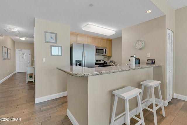 kitchen featuring appliances with stainless steel finishes, a breakfast bar area, kitchen peninsula, light stone countertops, and light brown cabinets