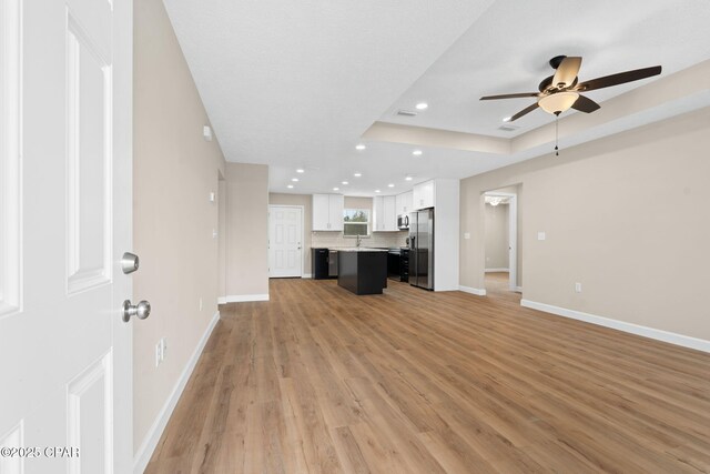 unfurnished living room featuring sink, a tray ceiling, ceiling fan, and light wood-type flooring