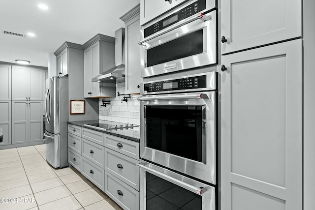 kitchen featuring wall chimney range hood, gray cabinetry, stainless steel appliances, light tile patterned floors, and dark stone counters