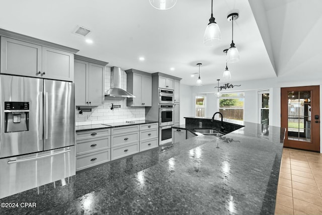 kitchen featuring appliances with stainless steel finishes, decorative light fixtures, dark stone counters, wall chimney exhaust hood, and sink