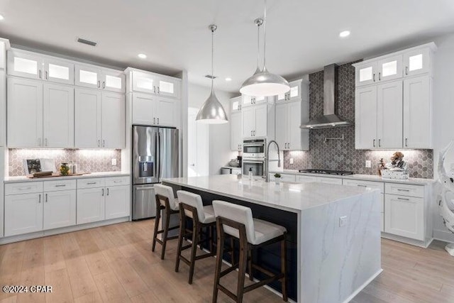 kitchen with white cabinetry, hanging light fixtures, stainless steel appliances, wall chimney range hood, and a kitchen island with sink