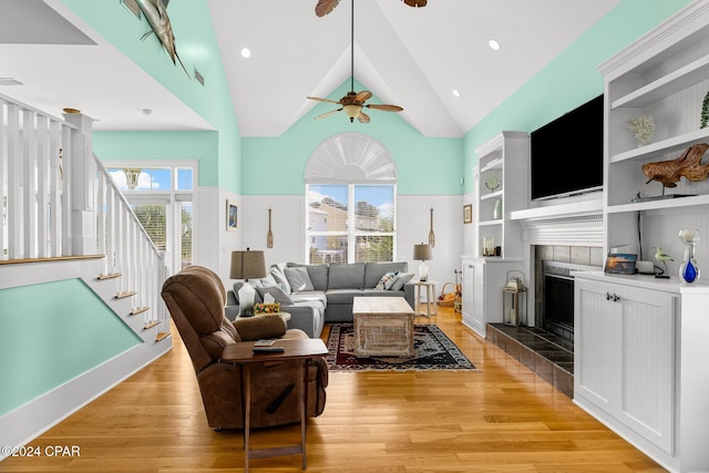 living room with high vaulted ceiling, ceiling fan, built in shelves, light hardwood / wood-style floors, and a tiled fireplace