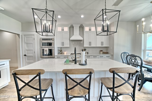 kitchen with premium range hood, stainless steel double oven, light wood-type flooring, an island with sink, and white cabinetry