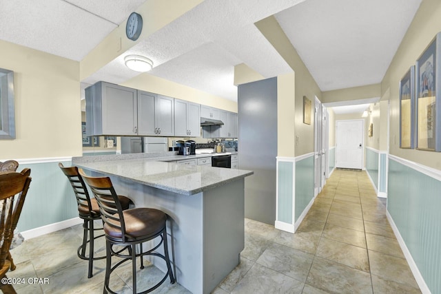 kitchen with electric range oven, a wainscoted wall, a kitchen breakfast bar, light stone countertops, and under cabinet range hood