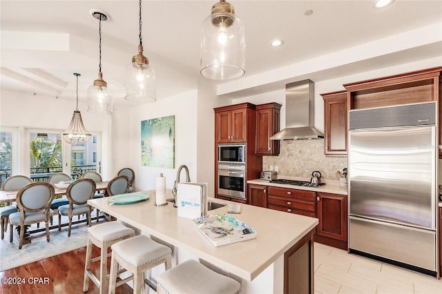 kitchen with wall chimney exhaust hood, sink, built in appliances, decorative light fixtures, and an island with sink