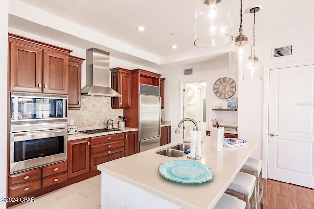 kitchen with pendant lighting, wall chimney range hood, sink, built in appliances, and light hardwood / wood-style floors