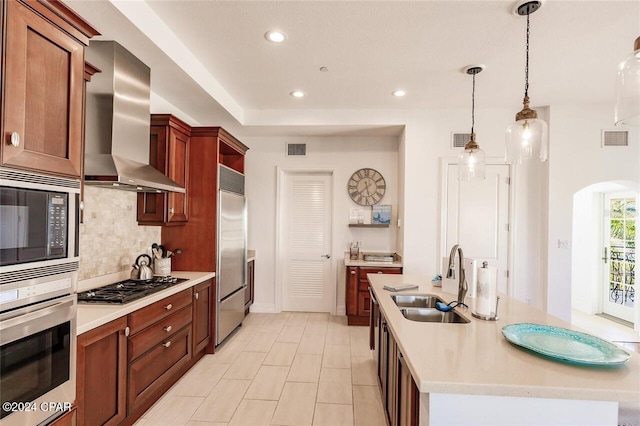 kitchen with a center island with sink, wall chimney range hood, sink, built in appliances, and decorative light fixtures