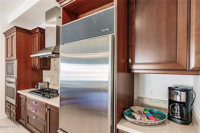 kitchen featuring built in appliances, light countertops, backsplash, wall chimney exhaust hood, and brown cabinetry