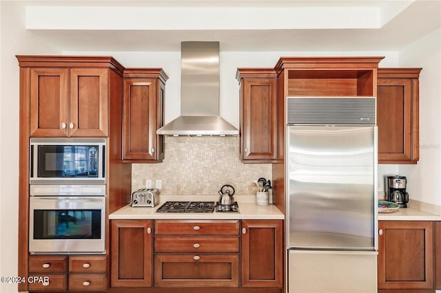 kitchen with brown cabinetry, decorative backsplash, built in appliances, light countertops, and wall chimney range hood