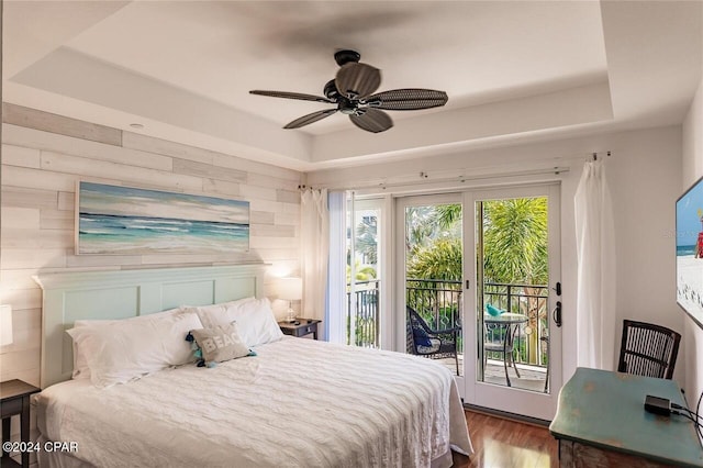 bedroom with access to exterior, ceiling fan, hardwood / wood-style floors, a tray ceiling, and wooden walls