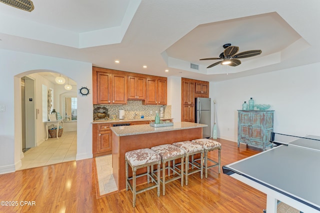 kitchen featuring arched walkways, backsplash, freestanding refrigerator, brown cabinetry, and a raised ceiling