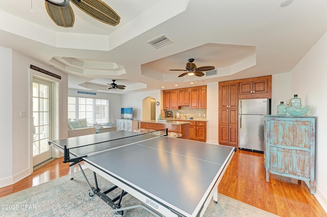 recreation room featuring light wood finished floors, a raised ceiling, visible vents, a ceiling fan, and baseboards